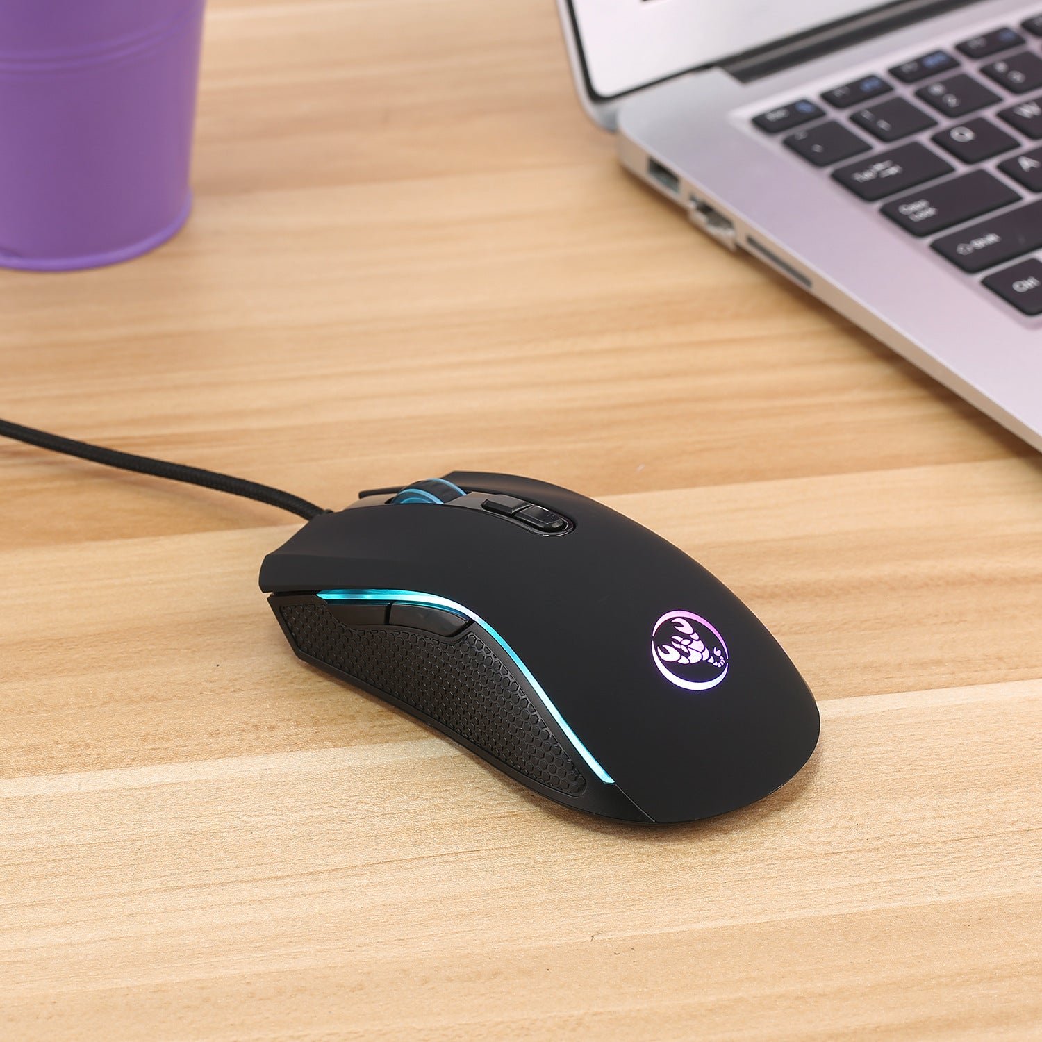 Black gaming mouse with RGB lighting on a wooden desk next to a laptop and purple cup.